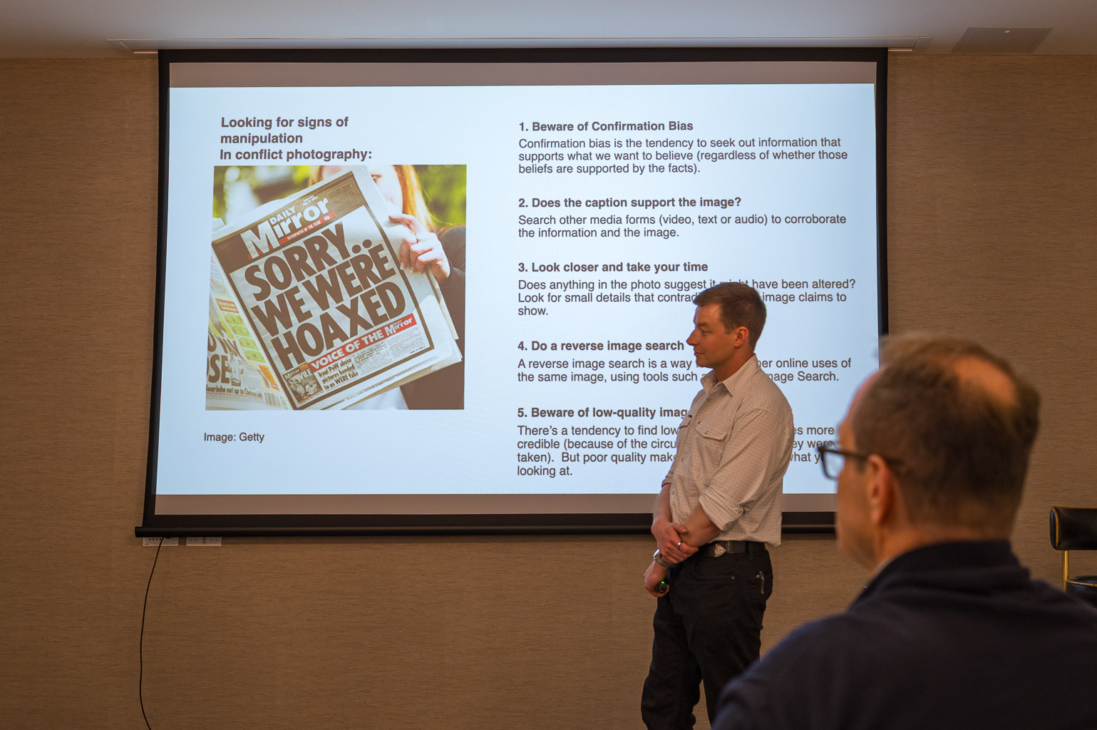 A presenter discusses signs of manipulation in conflict photography with a slide showing a newspaper headline reading 'Sorry We're Hoaxed' alongside bullet points outlining key concepts on critical photo analysis.