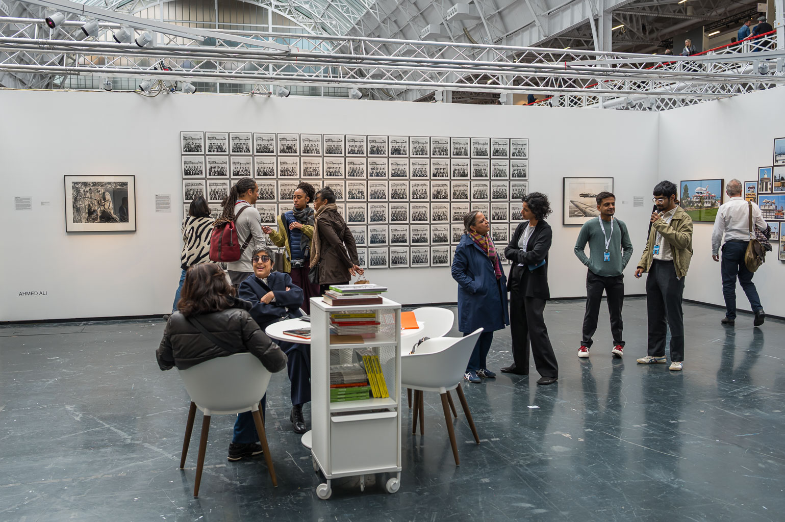 An art gallery scene featuring a group of people interacting in front of various photographs and artworks displayed on white walls. A small table with books is visible in the foreground, while visitors engage in conversation.