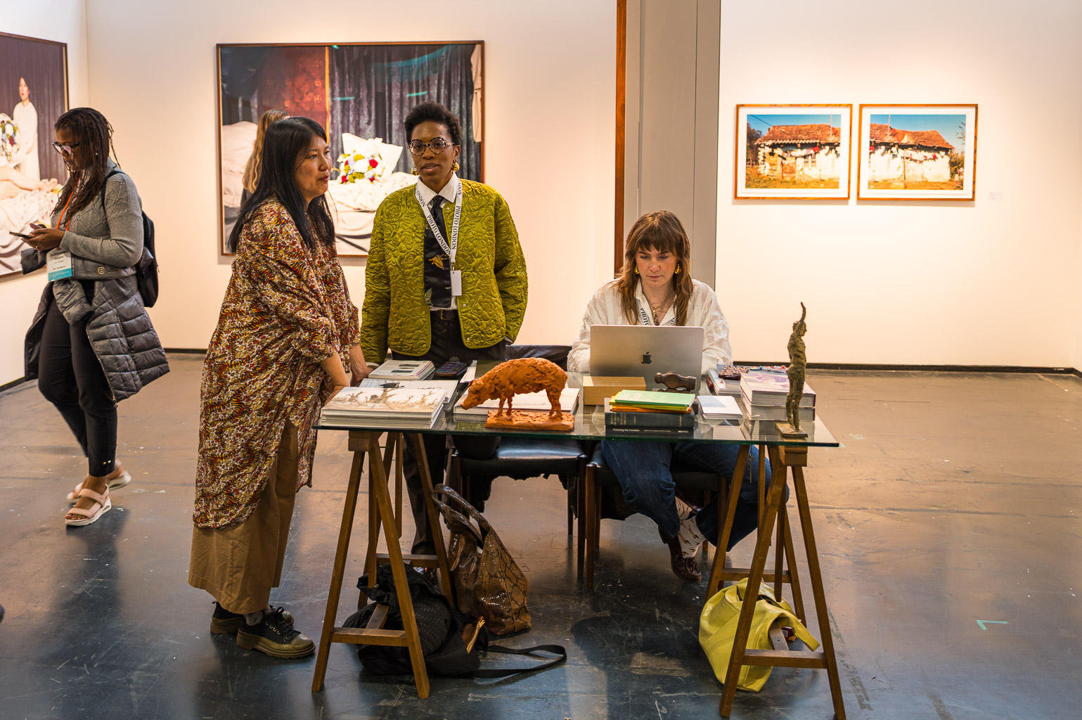 An art exhibition scene with three women. Two of them are conversing near a table displaying art books and sculptures, while a third woman is working on a laptop at the table. Various artworks are hanging on the walls in the background.