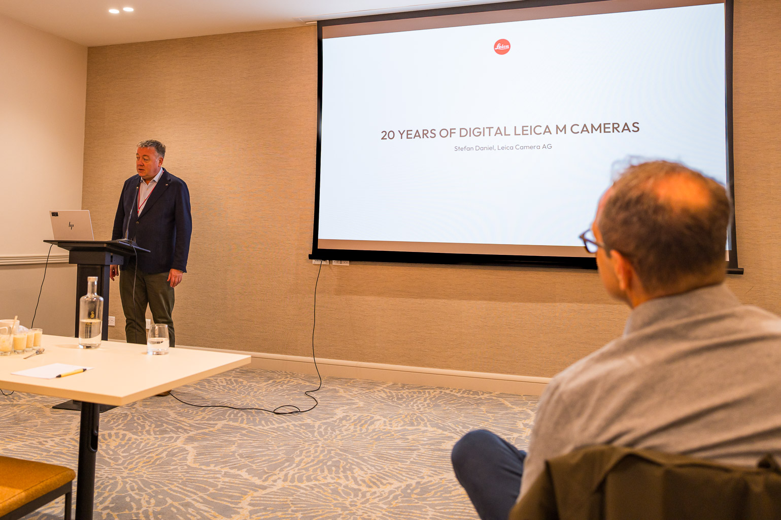 A presenter speaks at a conference about the topic '20 Years of Digital Leica M Cameras' while an audience member listens attentively.