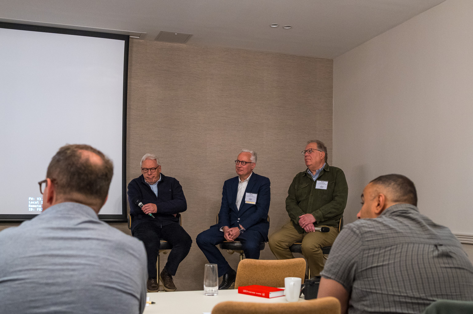 A panel discussion featuring three speakers seated on stools, with a projector screen in the background. Attendees are visible in the foreground, focusing on the panelists.