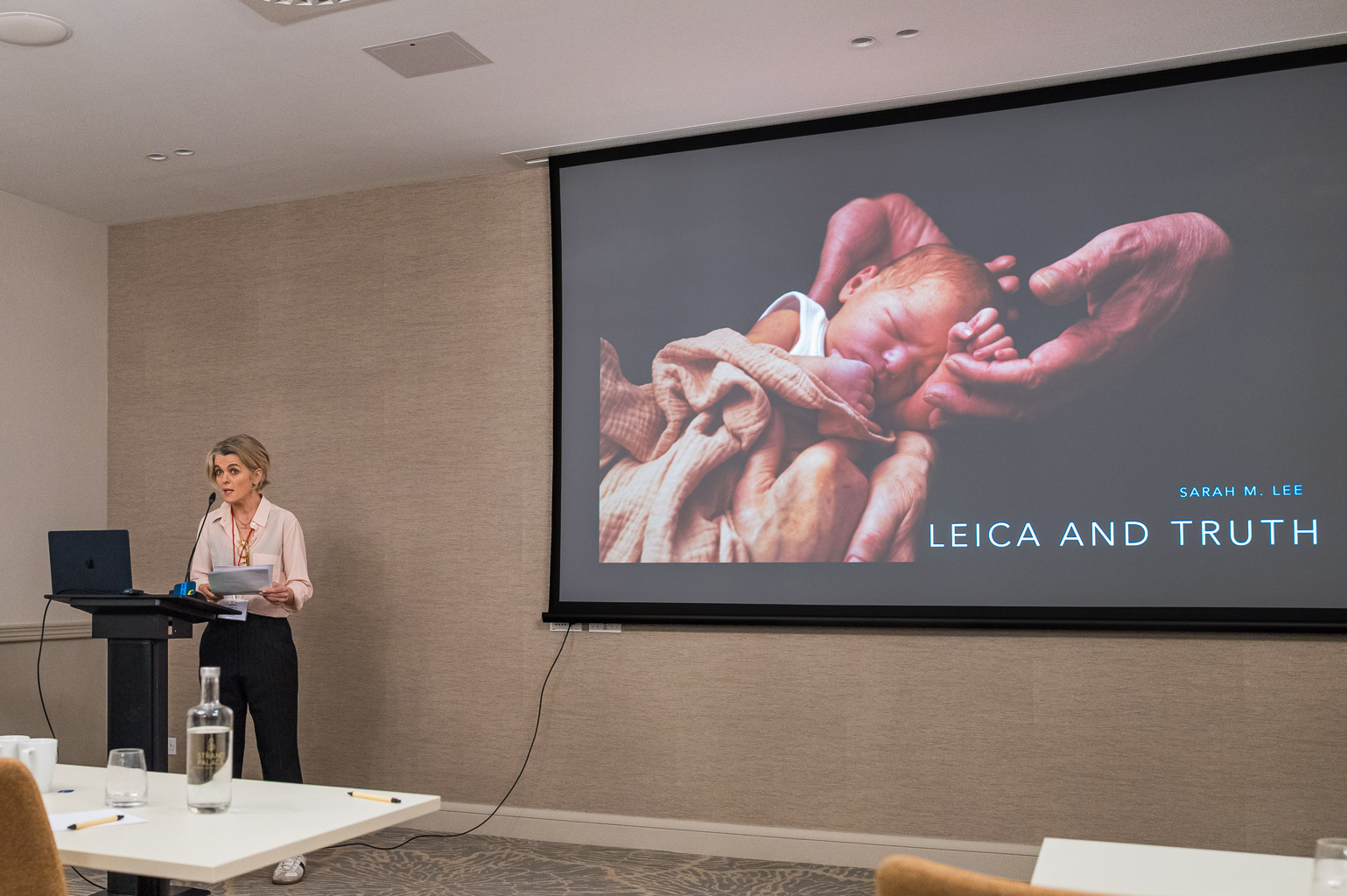 A speaker stands at a podium in a conference room, presenting on 'Leica and Truth' with a large screen behind displaying an image of a baby being held in hands. The speaker is wearing a light pink blouse and black pants.