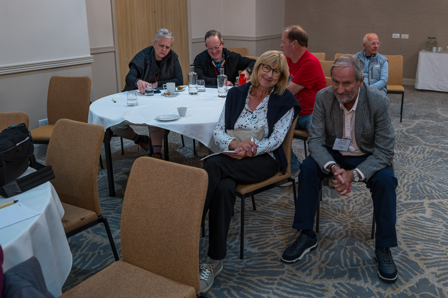 A group of six people sitting around a table in a conference room. Two people sit at the table, while four others are scattered around the room, engaged in conversation.