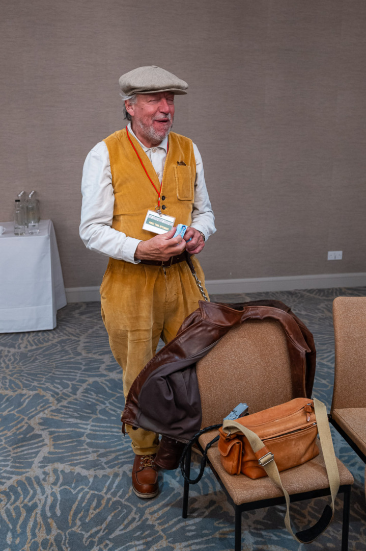 An elderly man wearing a flat cap and a yellow vest is standing, smiling, and holding a small object. He has a leather jacket draped over a chair and a brown messenger bag in his hand. The background shows a simple indoor setting.