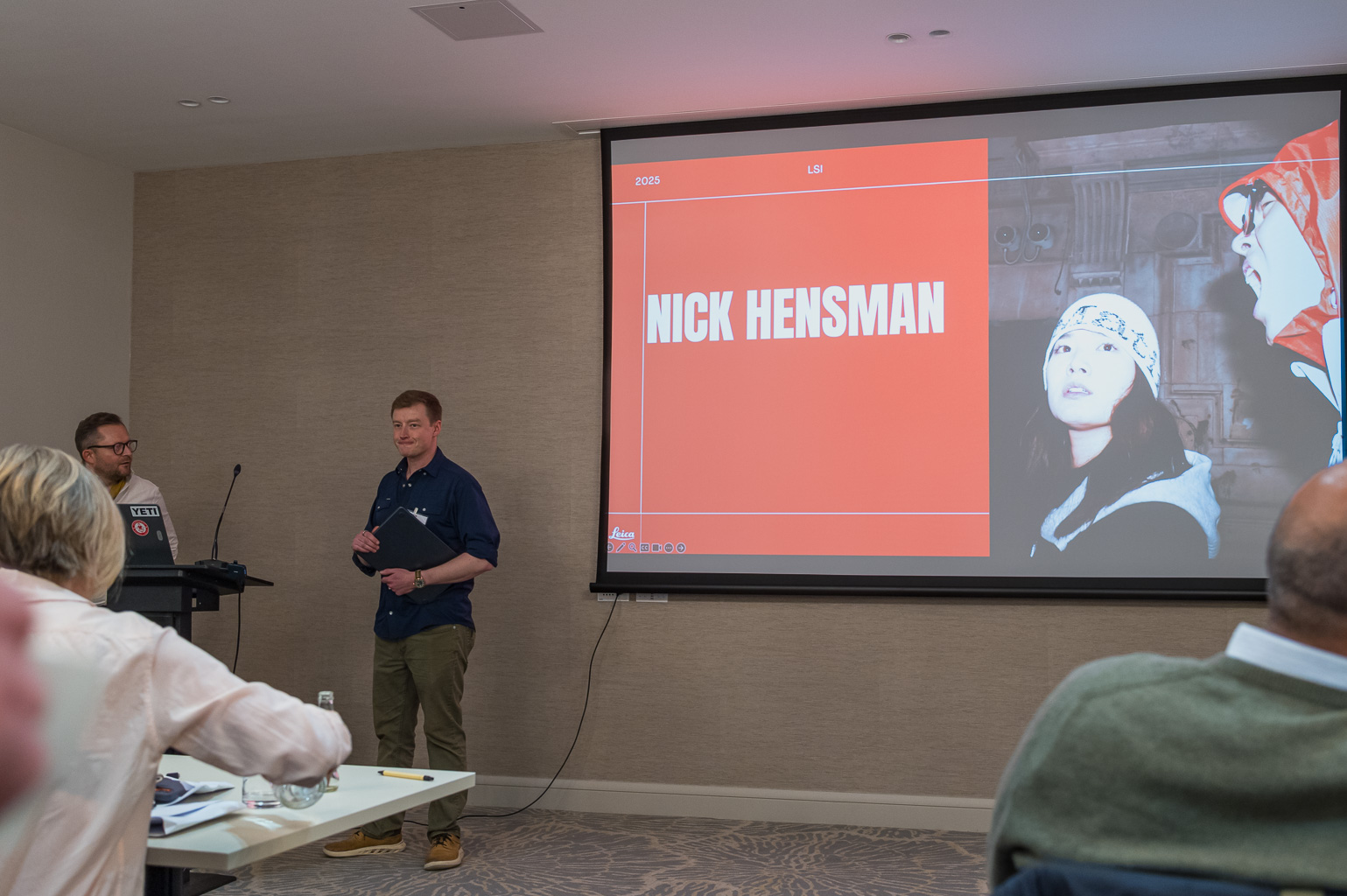 A speaker presenting in a conference room with a large screen displaying the name 'Nick Hensman.' An audience is visible, engaged in the presentation.