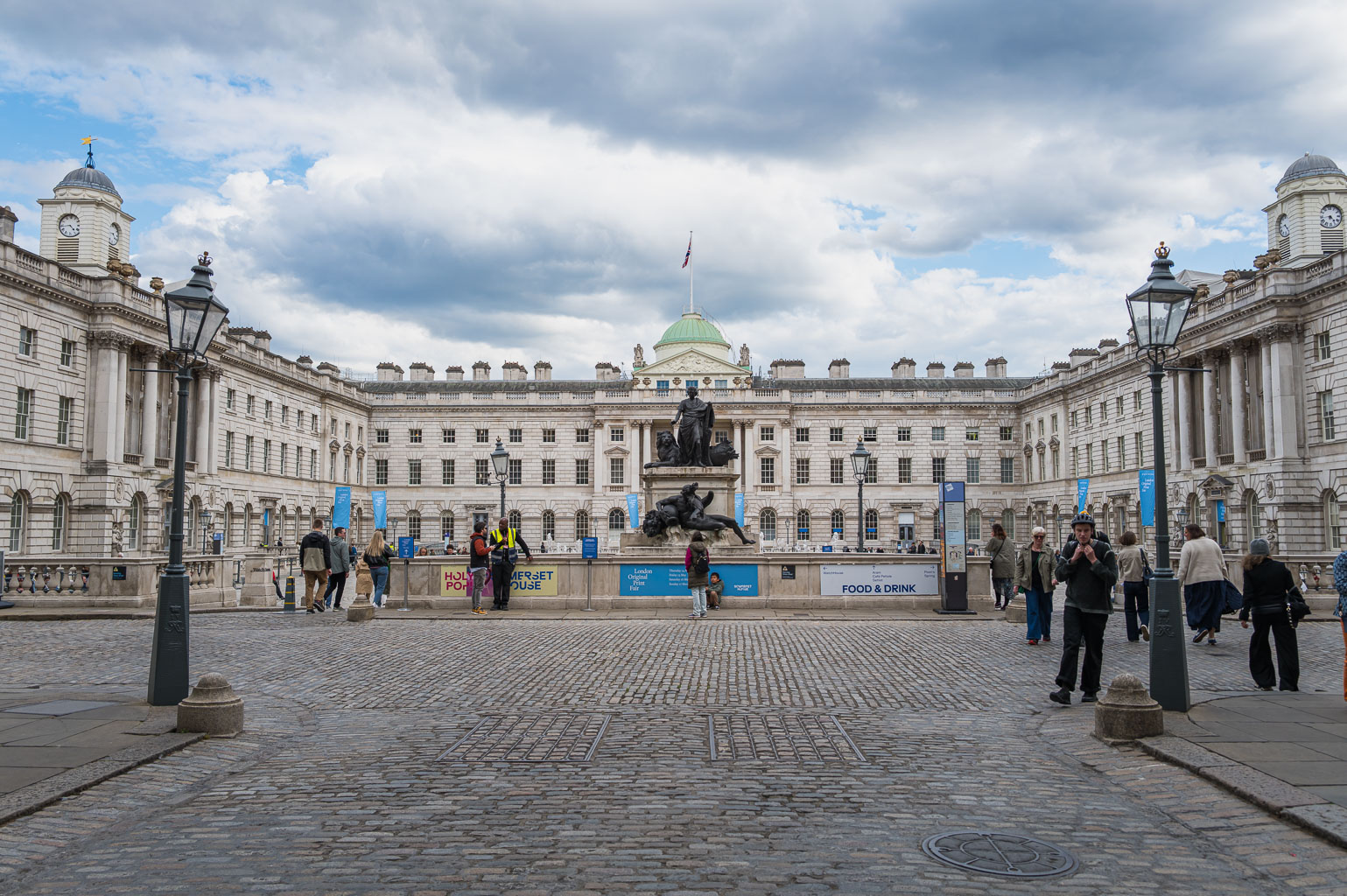 A view of Somerset House courtyard in London, featuring a statue, cobblestone pathway, and people interacting in a lively outdoor setting under a partly cloudy sky.