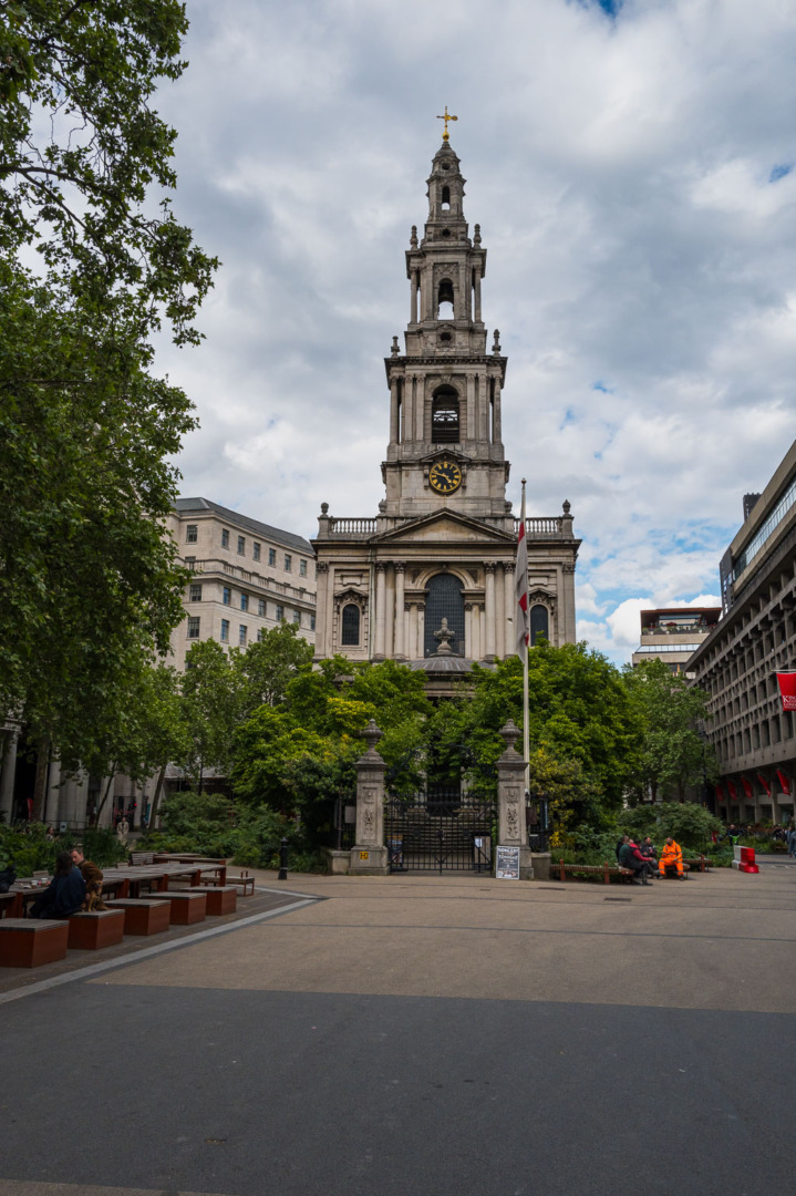 A historic church building with a tall clock tower, surrounded by trees and greenery in an urban setting. Benches and people are visible in the foreground, under a cloudy sky.