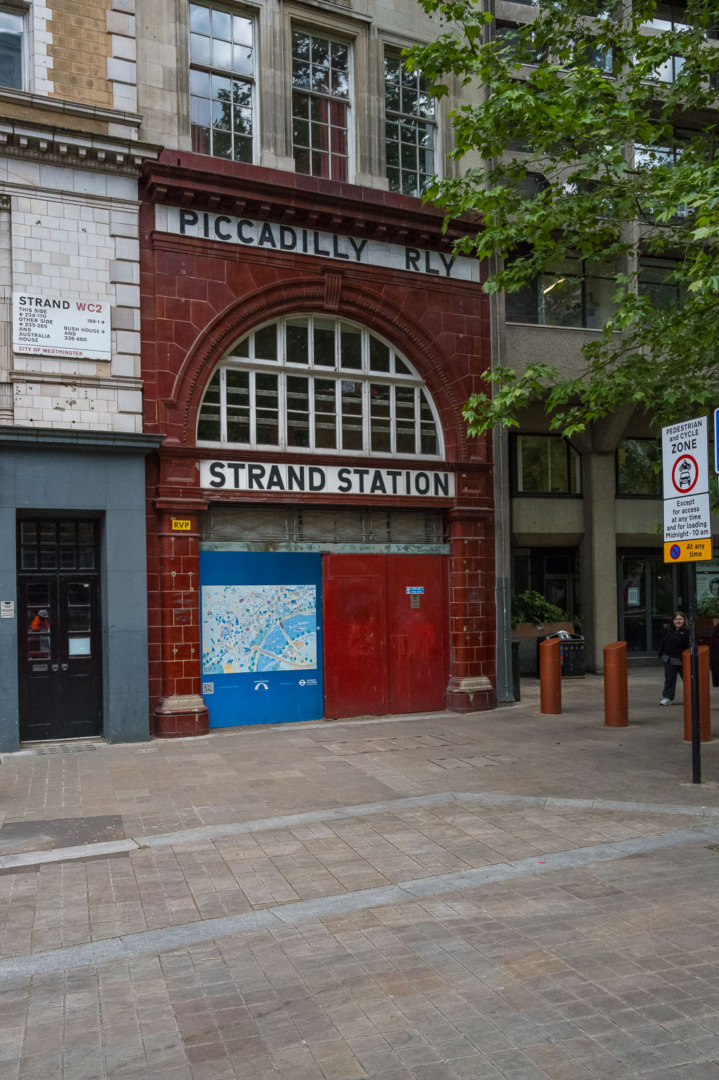 Exterior view of Strand Station with red brick archway and signage for Piccadilly Line, featuring a blue map display and nearby modern architecture.