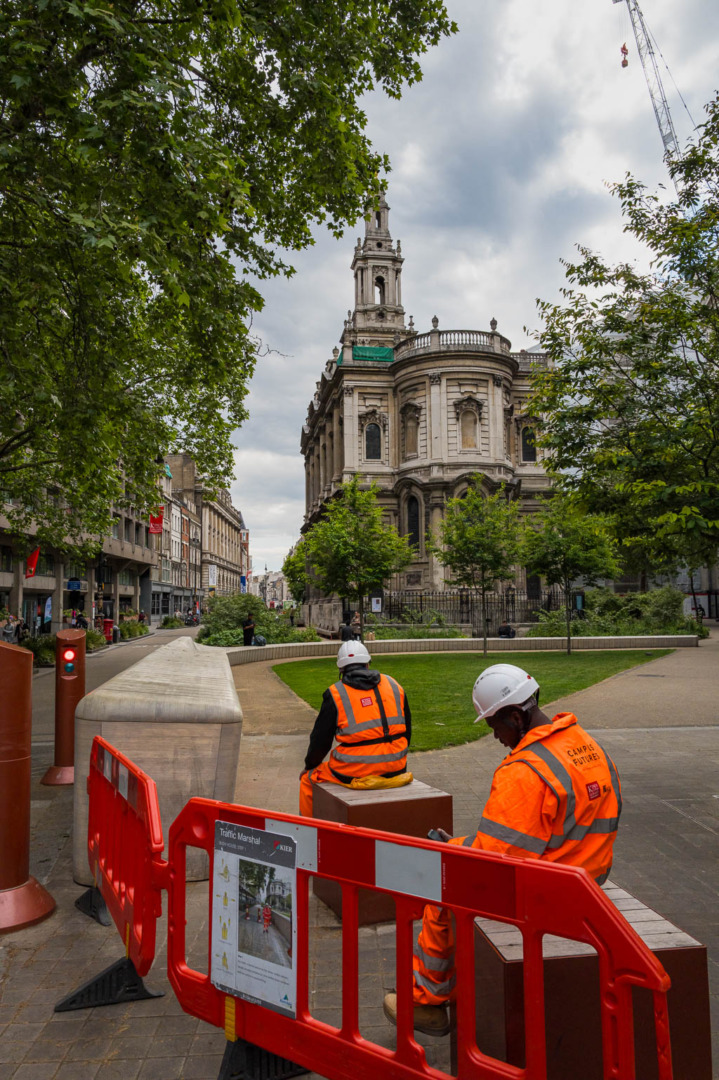 Two construction workers in safety gear sit near a traffic barrier in an urban setting, with a historic building in the background under cloudy skies.
