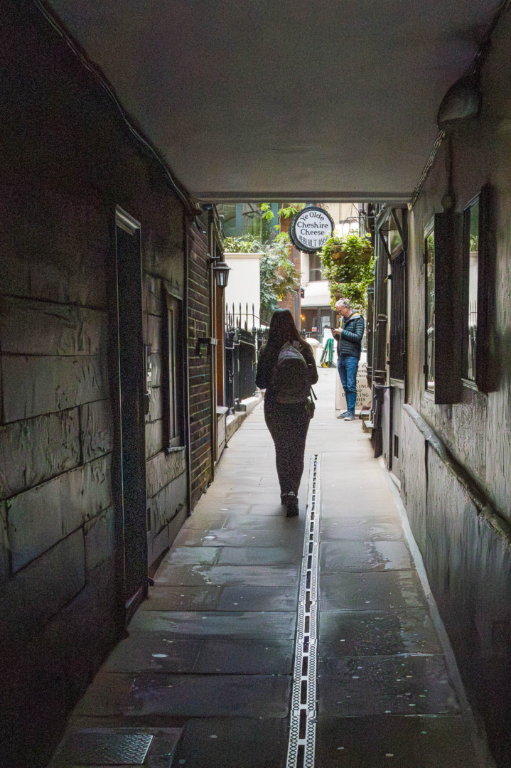 A narrow alleyway with textured walls, where a person with a backpack walks towards the camera. In the background, a man stands near a pub sign that reads 'The Olde Cheshire Cheese,' surrounded by greenery and a faintly lit path.