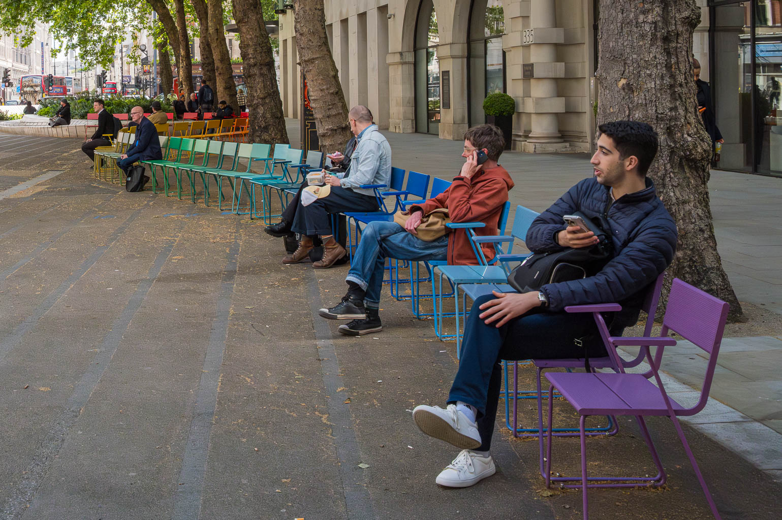 People sitting on colorful outdoor chairs in a public space, with trees and buildings in the background.
