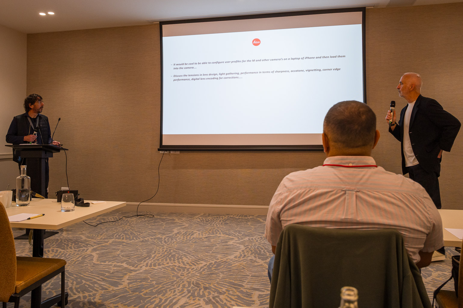 Two presenters speaking at a conference, one at a podium and the other standing beside a projection screen displaying text.