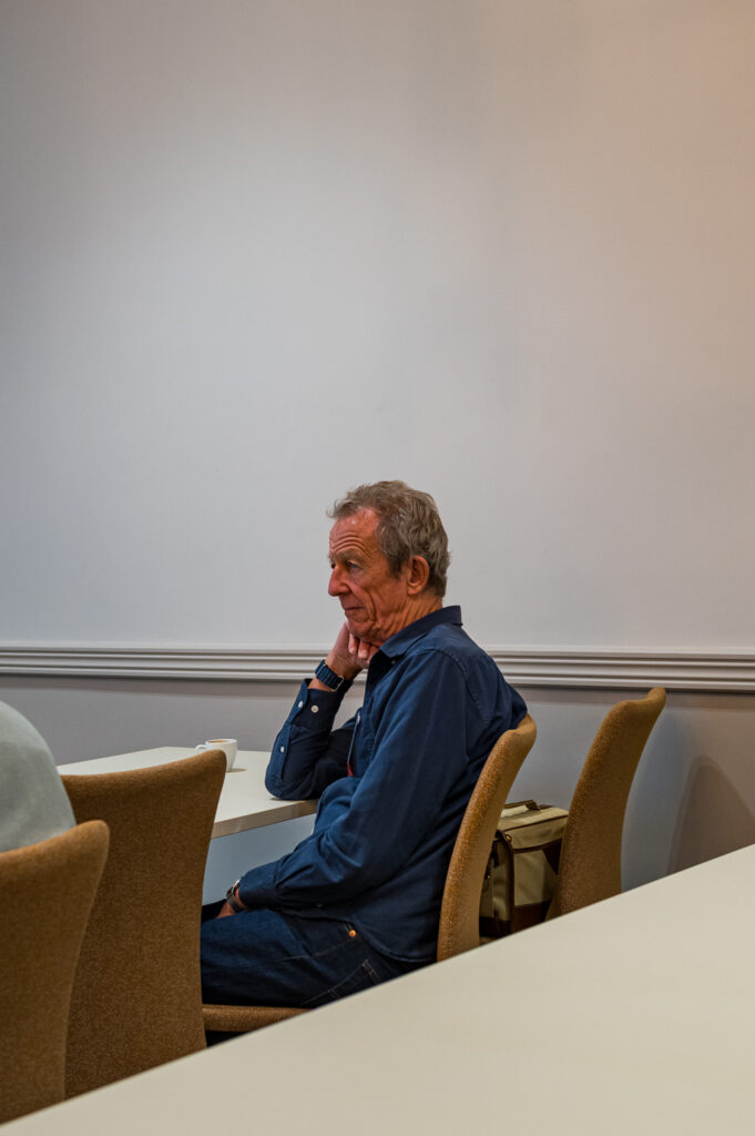 An older man sitting alone at a table in a meeting room, resting his chin on his hand, with a neutral background.