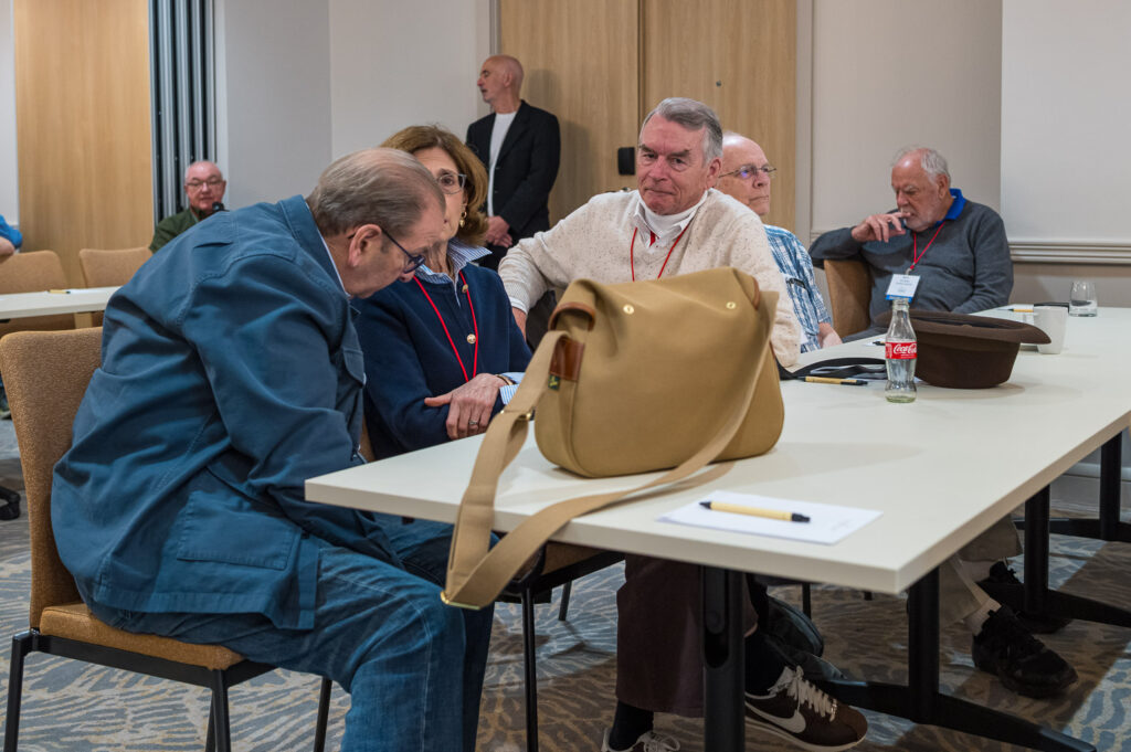 A group of older adults seated at a table in a conference room, engaged in conversation. One individual leans towards another, while others look on. Personal items like a bag and a soda can are visible on the table.