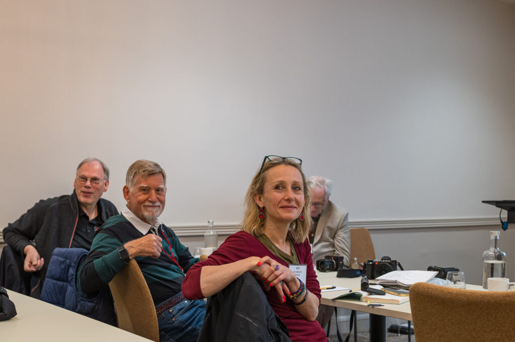 A group of four people engaged in a meeting, with one woman smiling at the camera while sitting at a table, and three men visible in the background, two of whom are also looking towards the camera.