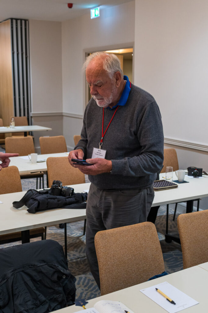 An older man in a gray sweater and blue collared shirt stands in a classroom-like setting, looking at his phone. He wears a name tag and is surrounded by empty tables with chairs and personal items, including a camera and notebooks.