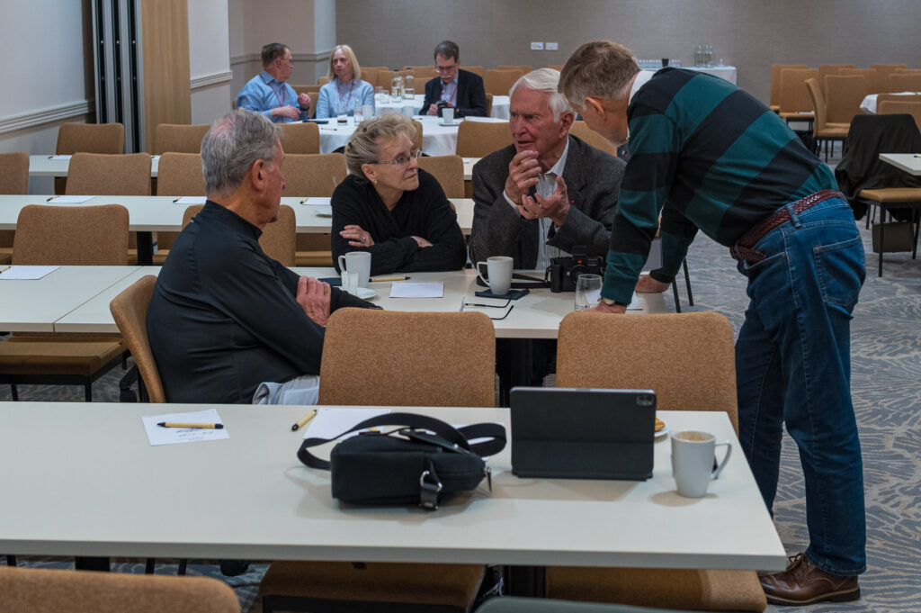 A group of four people engaged in conversation at a meeting table in a conference room, with additional participants in the background. Coffee cups and notepads are on the tables.