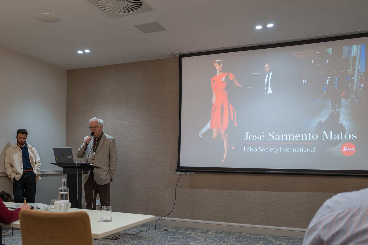 A speaker, wearing glasses and a blazer, presents at a podium while another man listens nearby. The presentation behind them features a slide with text reading 'José Sarmento Matos, Leica Society International' alongside images of people in formal and casual attire.