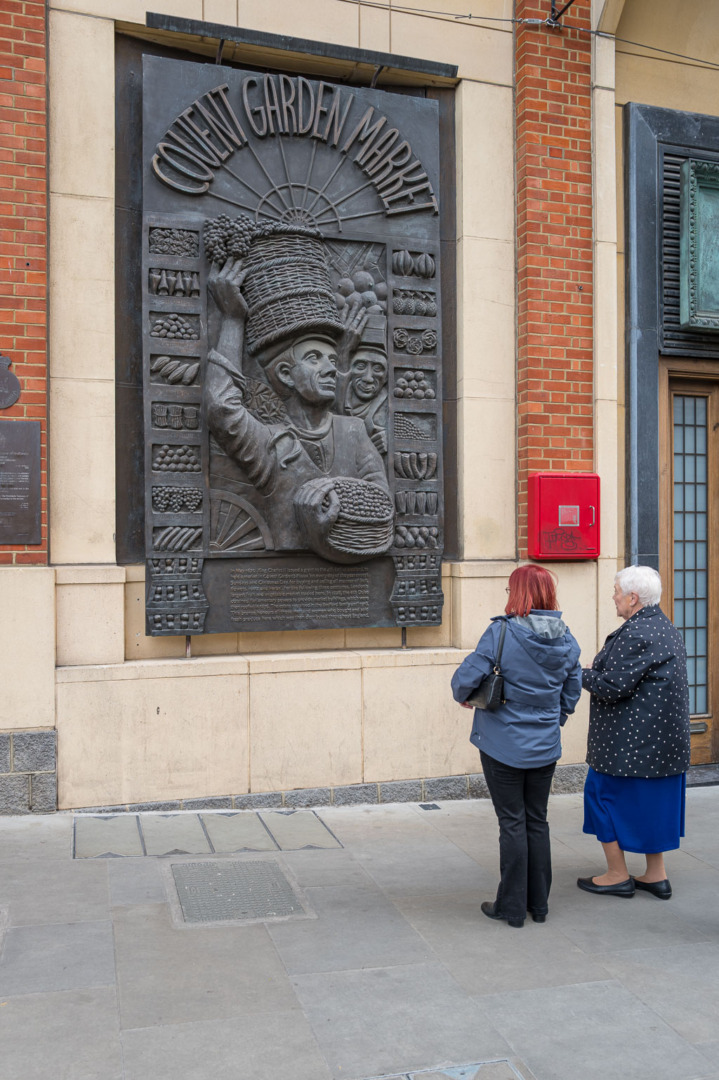 Two women examining a bronze relief depicting scenes from Covent Garden Market, featuring market workers and produce, mounted on a brick wall.