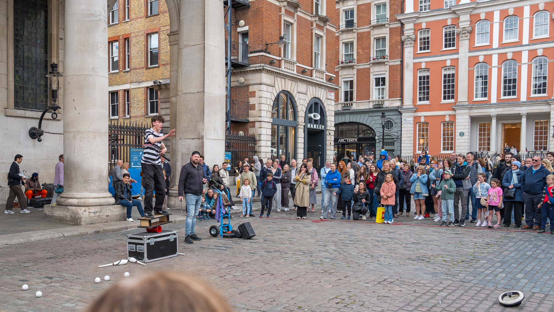 A street performer entertains a large crowd in a plaza, standing on a box and gesturing while juggling objects, with spectators of various ages watching attentively.