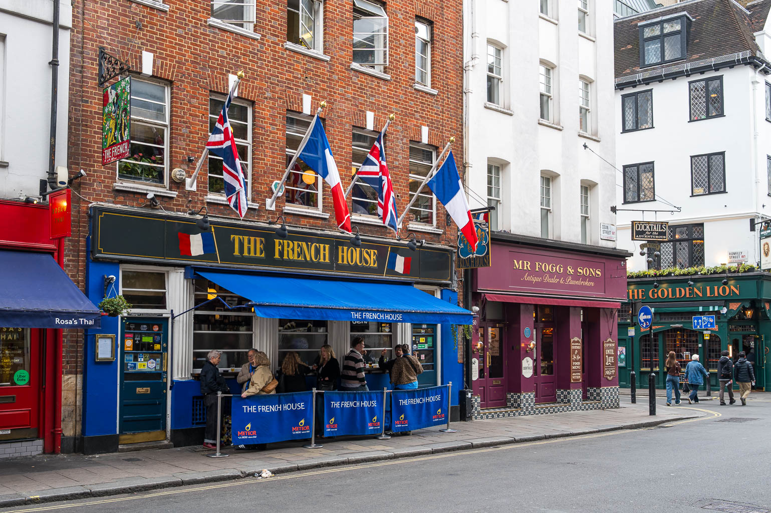 A street view featuring the French House pub with British and French flags, surrounded by other themed establishments.