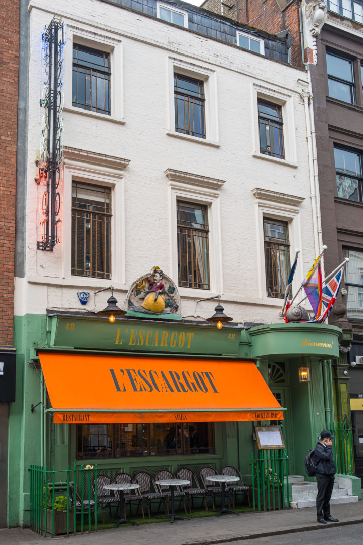Exterior view of the L'Escargot restaurant, featuring an orange awning with the restaurant name, decorative features above the entrance, and outdoor seating.