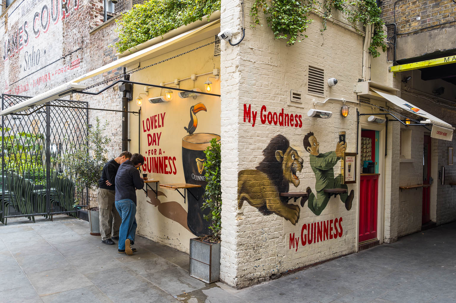 A cozy outdoor pub setting with two people enjoying drinks at a bar. The wall features a colorful mural advertising Guinness, depicting a cheerful toucan and a playful scene with a lion and a man. Lush greenery and string lights create a welcoming ambiance.