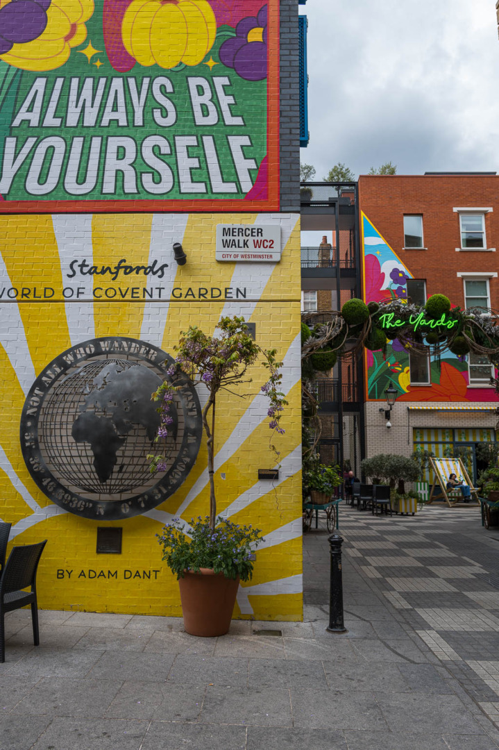 A vibrant mural featuring the phrase 'Always Be Yourself' with colorful designs, located on a wall near Stanfords in Covent Garden. The mural includes a globe illustration and is part of a lively shopping area with outdoor seating and planters.