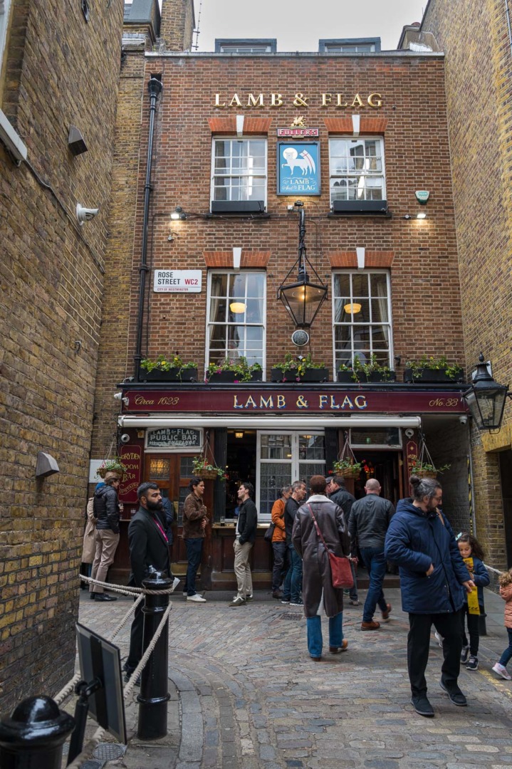 Exterior of the Lamb & Flag pub with patrons gathered outside, featuring brick walls and a sign above the entrance.