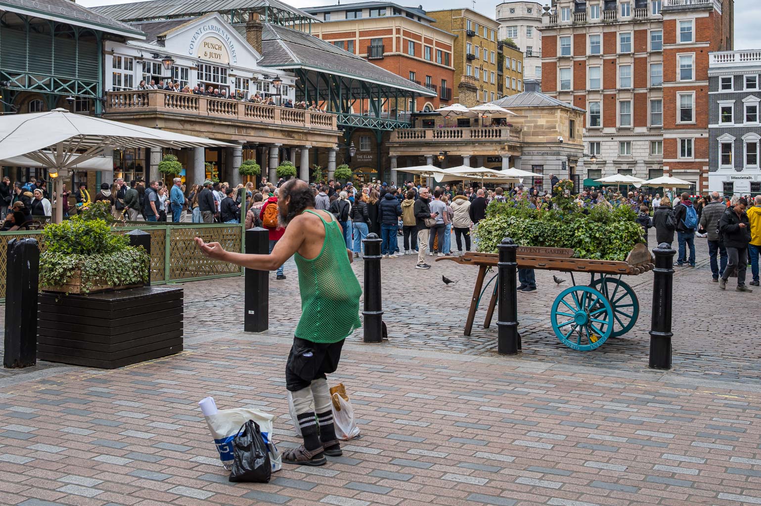 A street performer in a green shirt entertaining a crowd in Covent Garden, London, with people gathered around and buildings in the background.