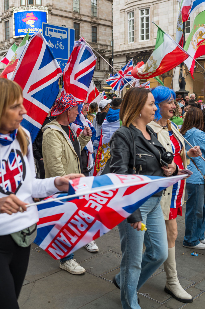 A crowd of people celebrating, holding British flags while walking on a street with buildings in the background.