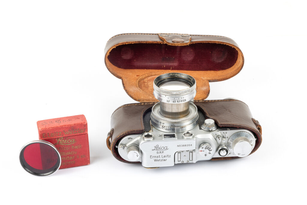 A vintage Leica camera displayed with its leather case, a lens, and a red filter.