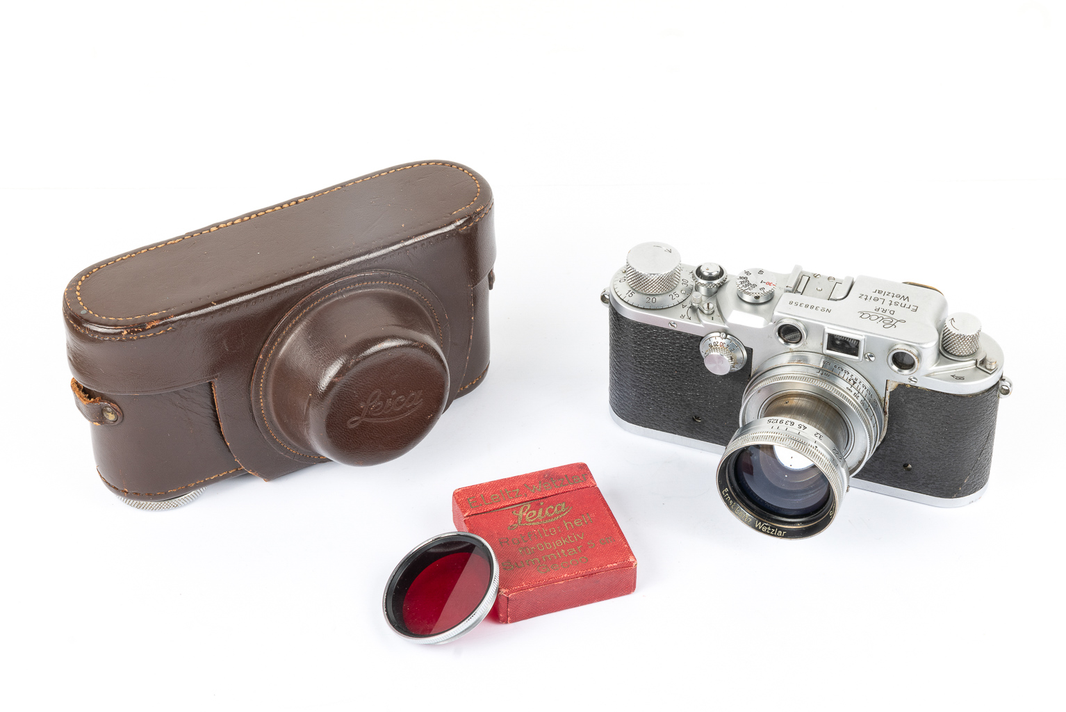 A vintage Leica camera with a silver body and a lens, accompanied by a brown leather case and a red filter box.