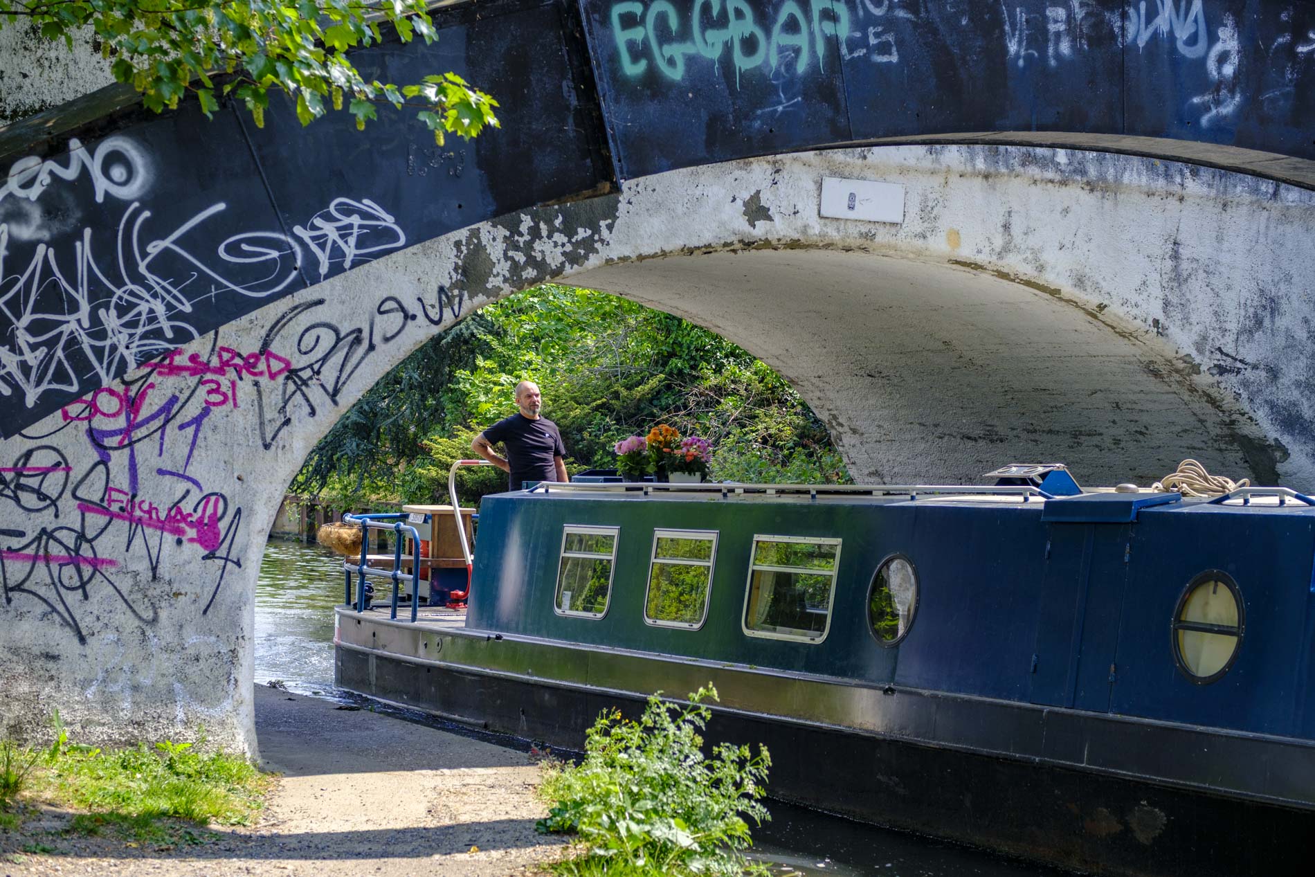 A man stands on a narrowboat near a canal under a graffiti-covered bridge, surrounded by lush greenery.