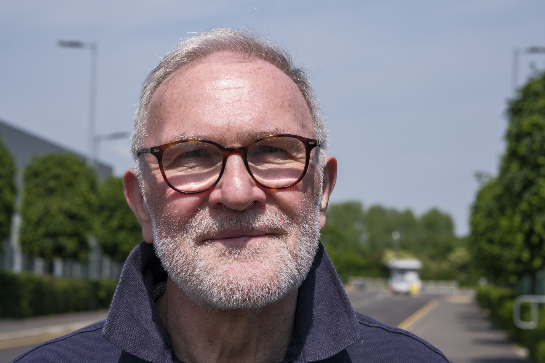 A smiling older man with glasses and a grey beard, standing outdoors on a sunny day, with trees and a road visible in the background.
