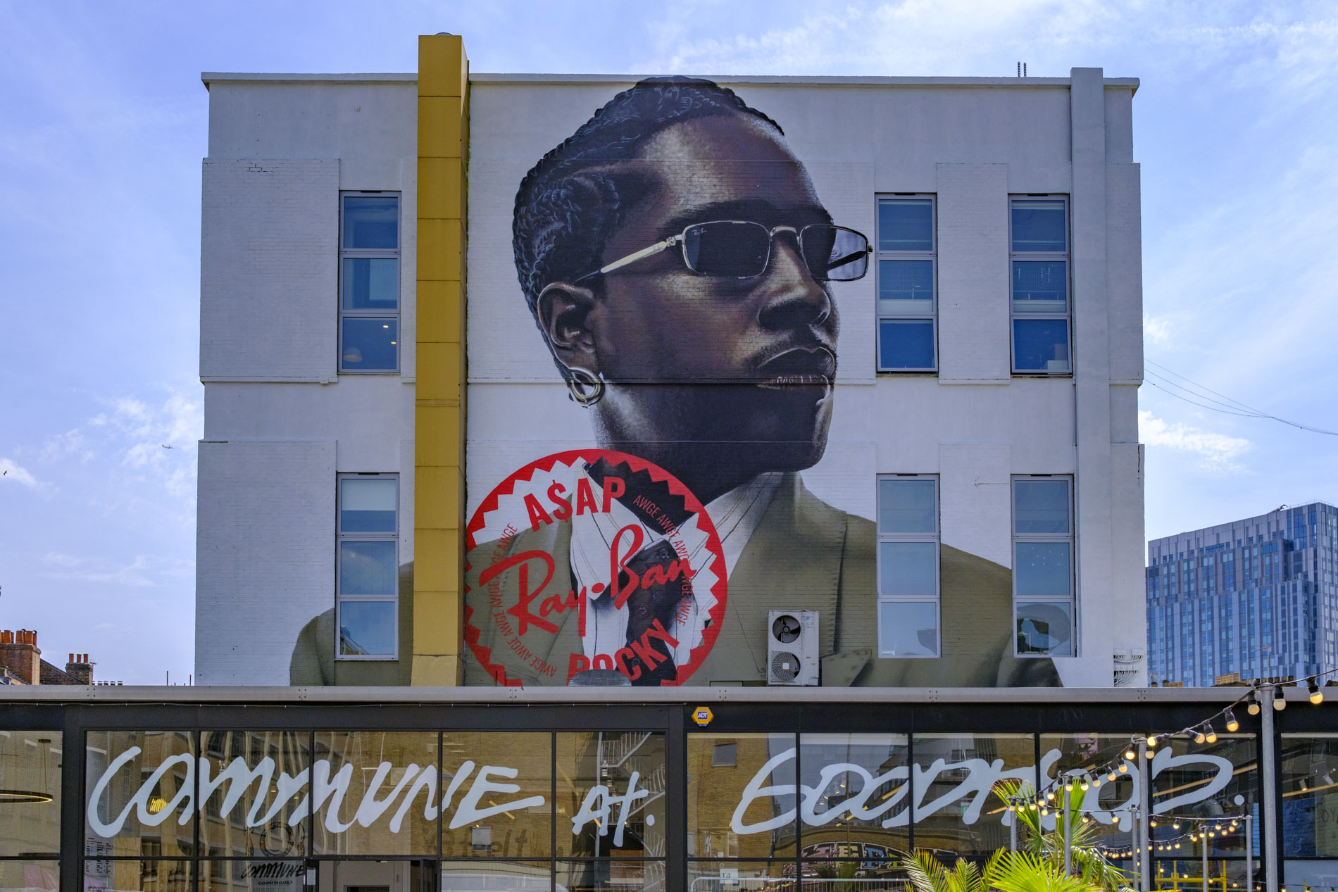Large mural of a man wearing sunglasses on the side of a building, featuring the text 'ASAP' and 'Ray-Ban' in a circular design beneath the artwork.
