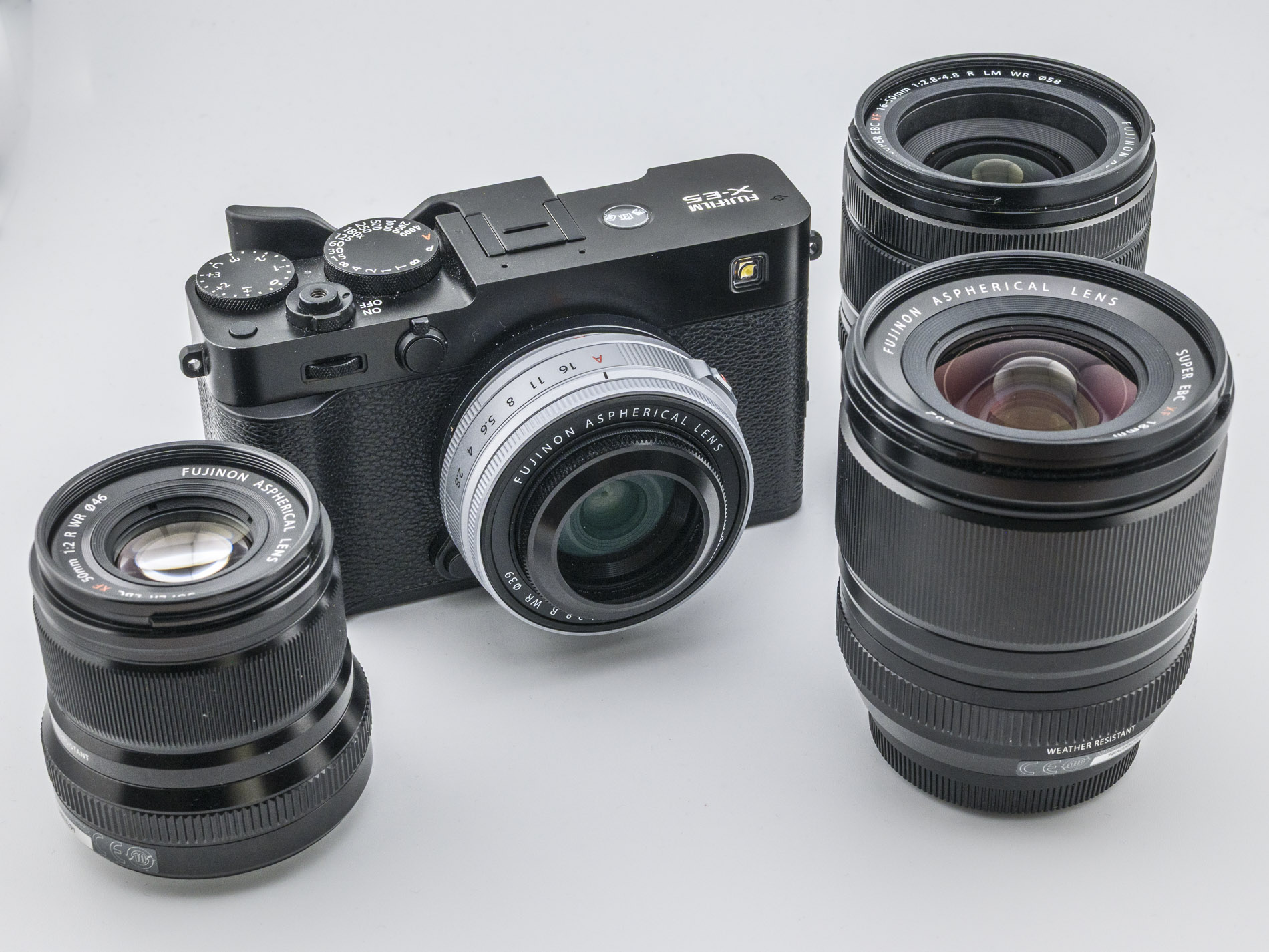 A Fujifilm X-Series camera with four interchangeable lenses positioned around it on a light background.