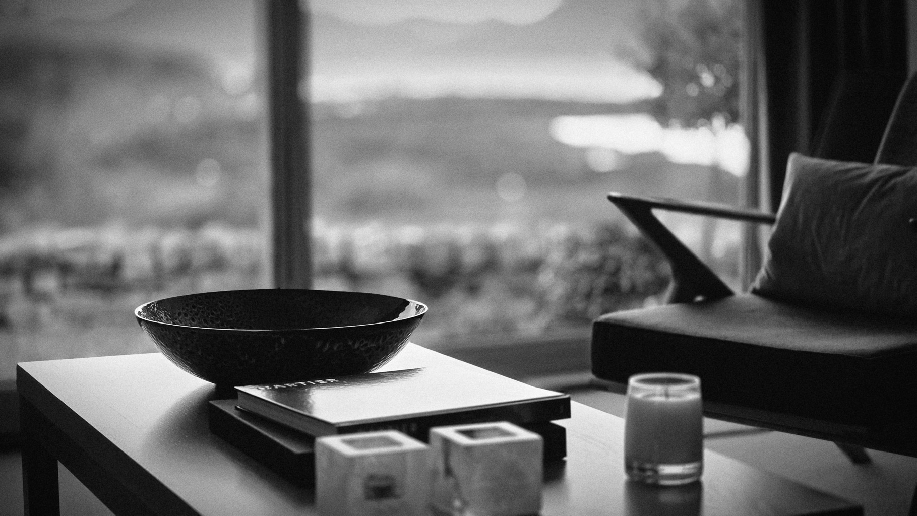 A cozy living space with a black bowl atop a wooden table, stacks of books, a lit candle, and a stylish chair visible in the background through a large window, offering a serene view.