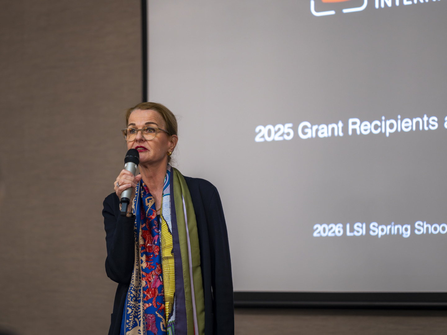 A woman with glasses speaks into a microphone at a presentation, wearing a colorful scarf and a dark jacket, with a screen behind her displaying the text '2025 Grant Recipients and 2026 LSI Spring Shoots'.