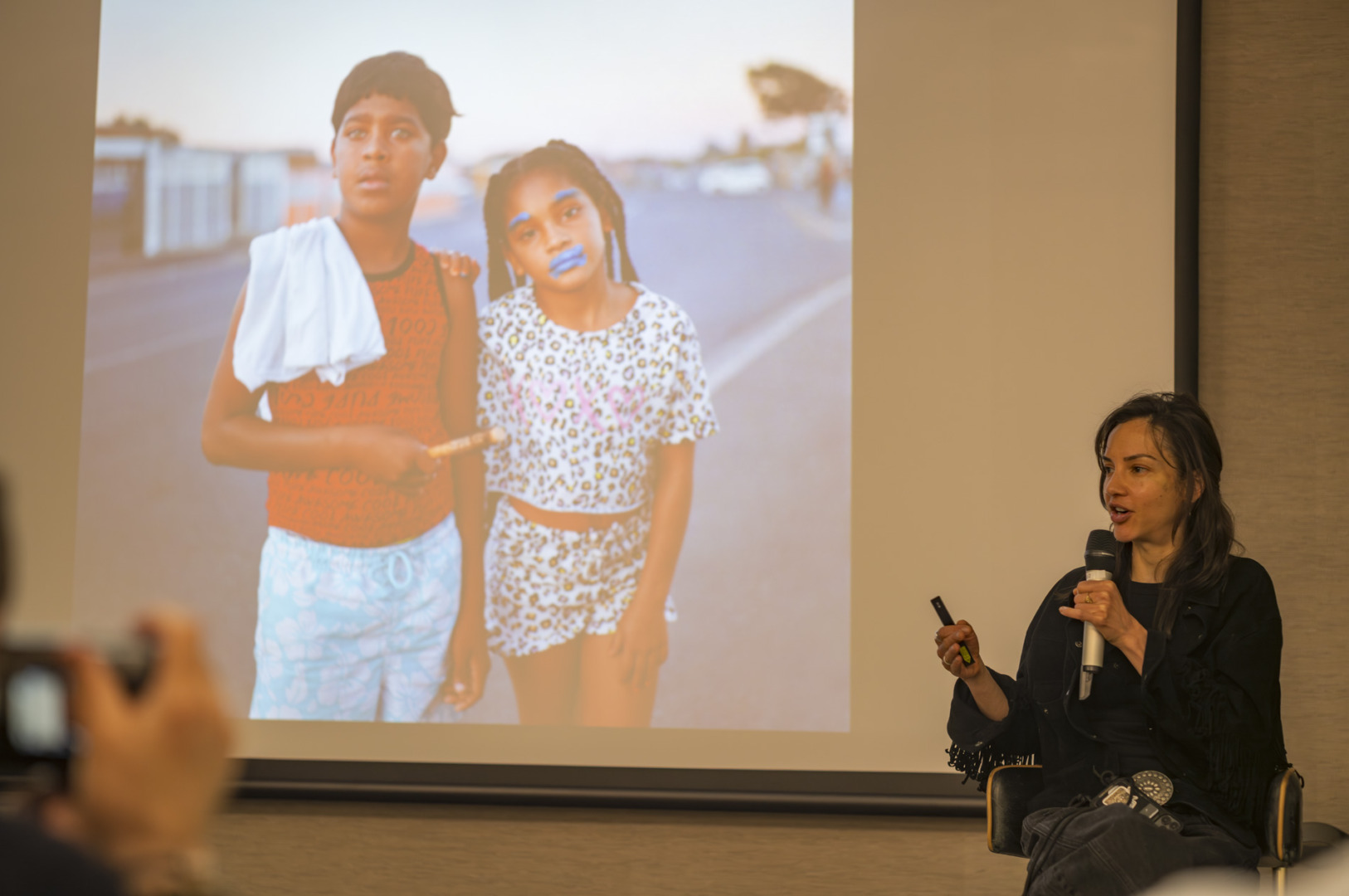 A speaker presents in front of a large screen displaying an image of two young girls, one with a towel over their shoulder, and the other with blue face paint.