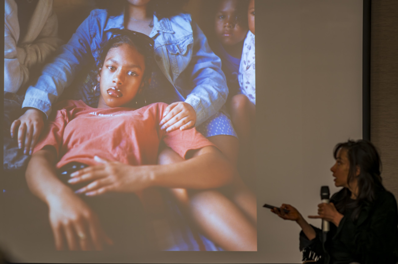 A person giving a presentation beside a large image of a girl lying down, looking contemplative, surrounded by others. The setting appears to be an indoor event.