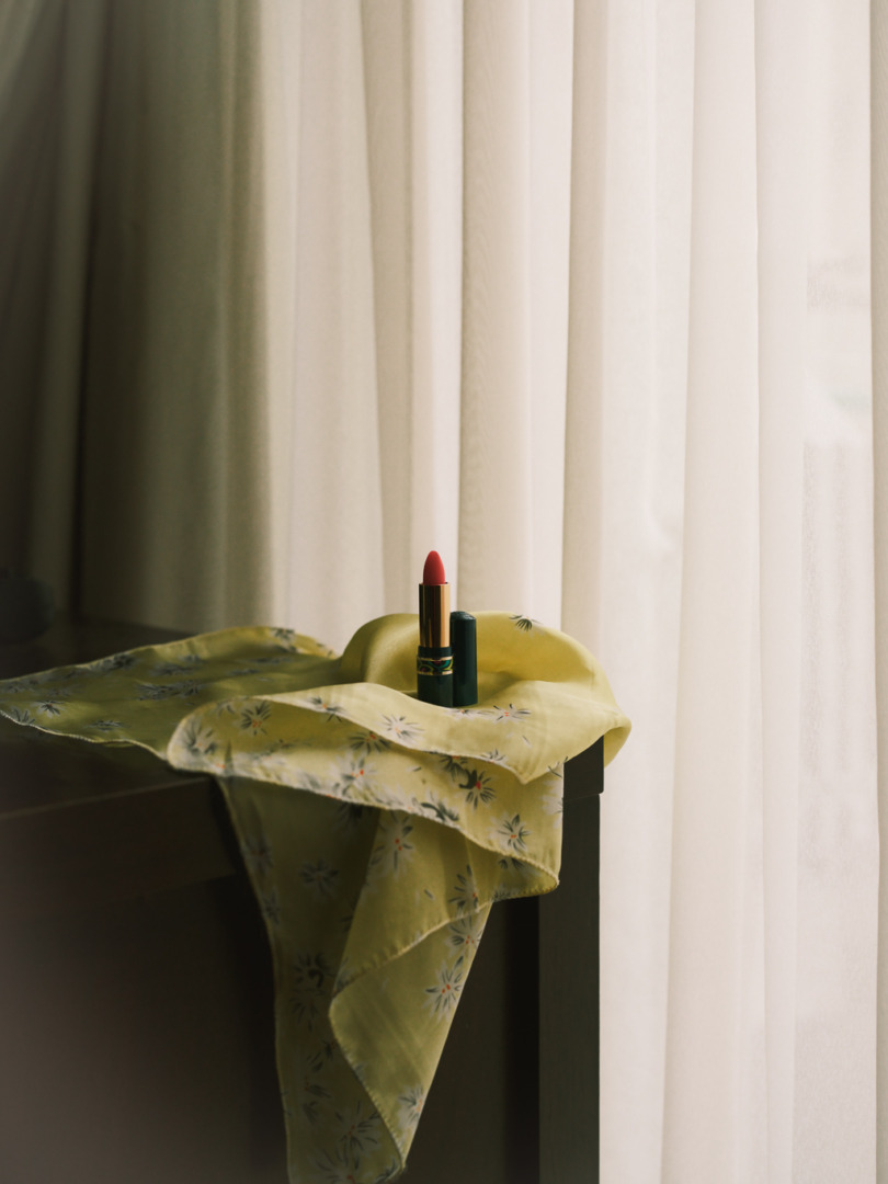 A bright red lipstick and a green lipstick tube placed on a yellow floral scarf, with soft curtains in the background.