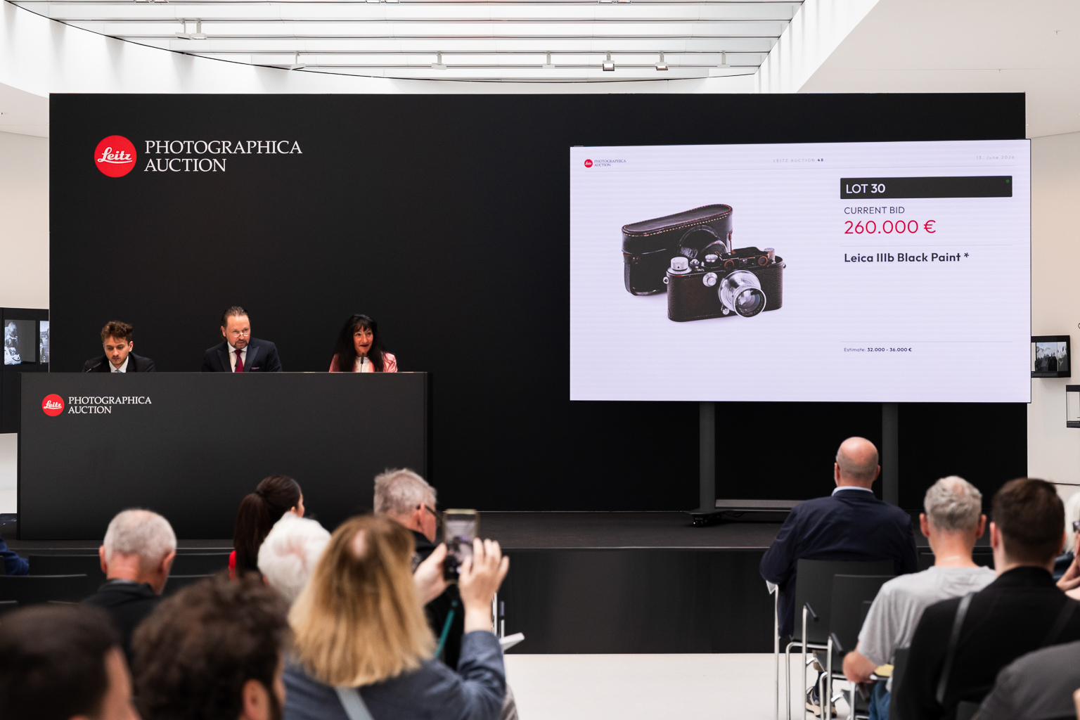 A live auction scene at the Leitz Photographic Auction featuring an item display with a current bid of 260,000 € for a Leica IIIb Black Paint camera. Three auctioneers are seated at a podium, while attendees view the auction.