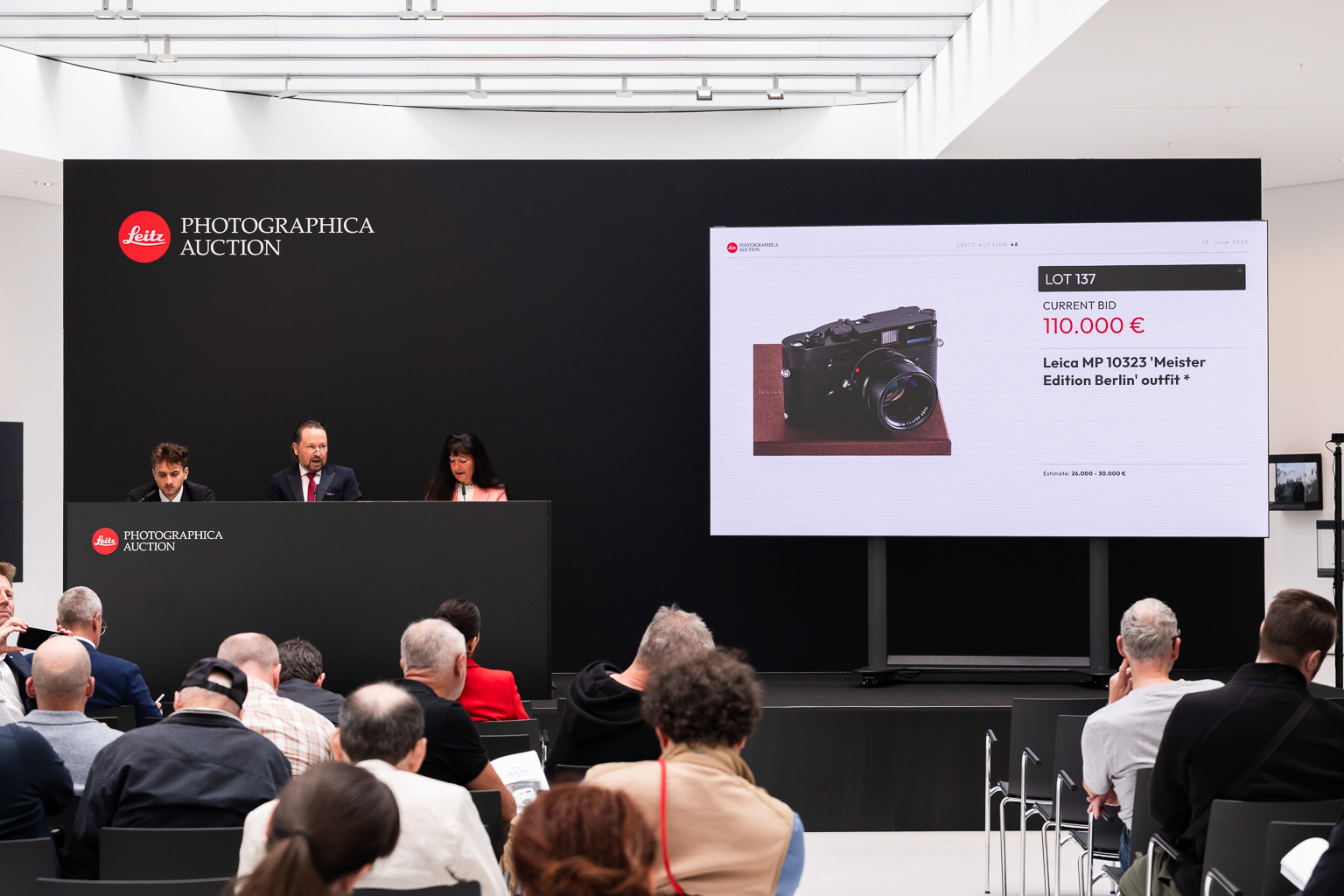 A live auction event at Leitz, featuring a bidding session for a Leica MP 10323 'Meister Edition Berlin' outfit, with a current bid of 110,000 euros displayed on a screen.