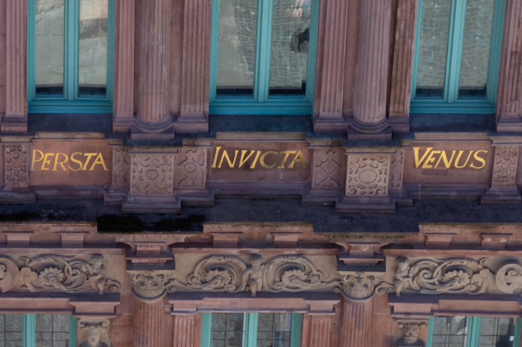 Close-up of ornate architectural detail featuring the words 'PERSTA INVICTA VENUS' inscribed in gold on a building facade, surrounded by intricate stone carvings and turquoise windows.