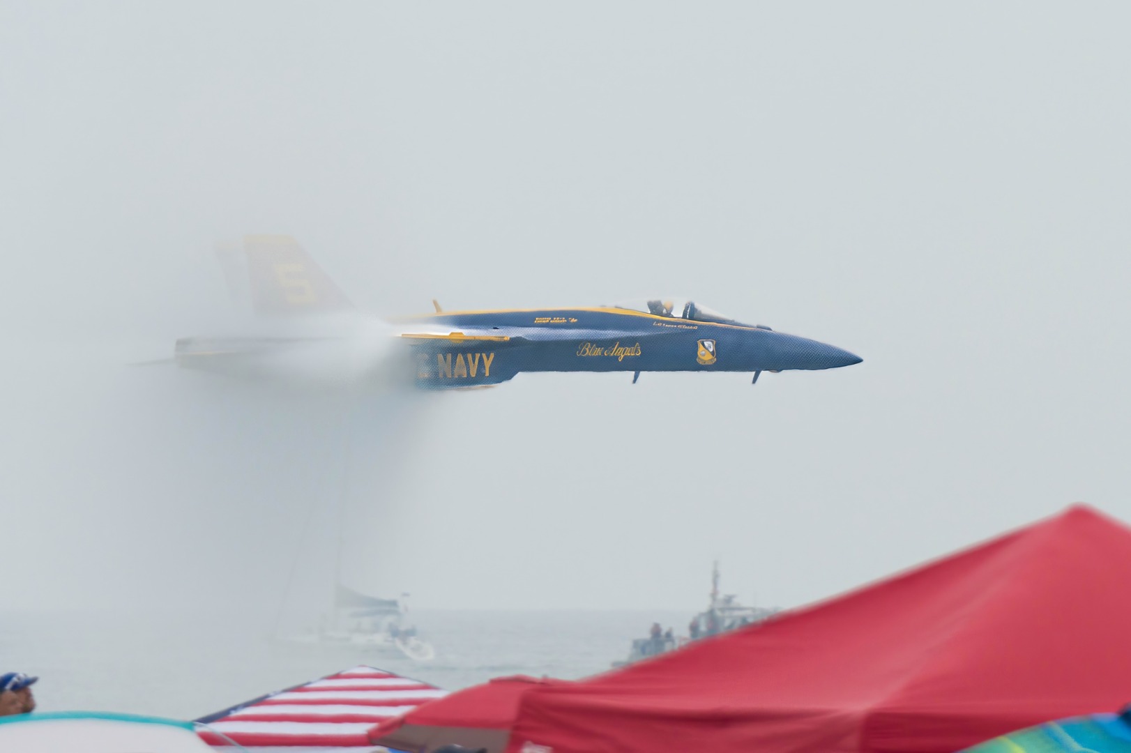 Blue fighter jet flying low above beach tents.
