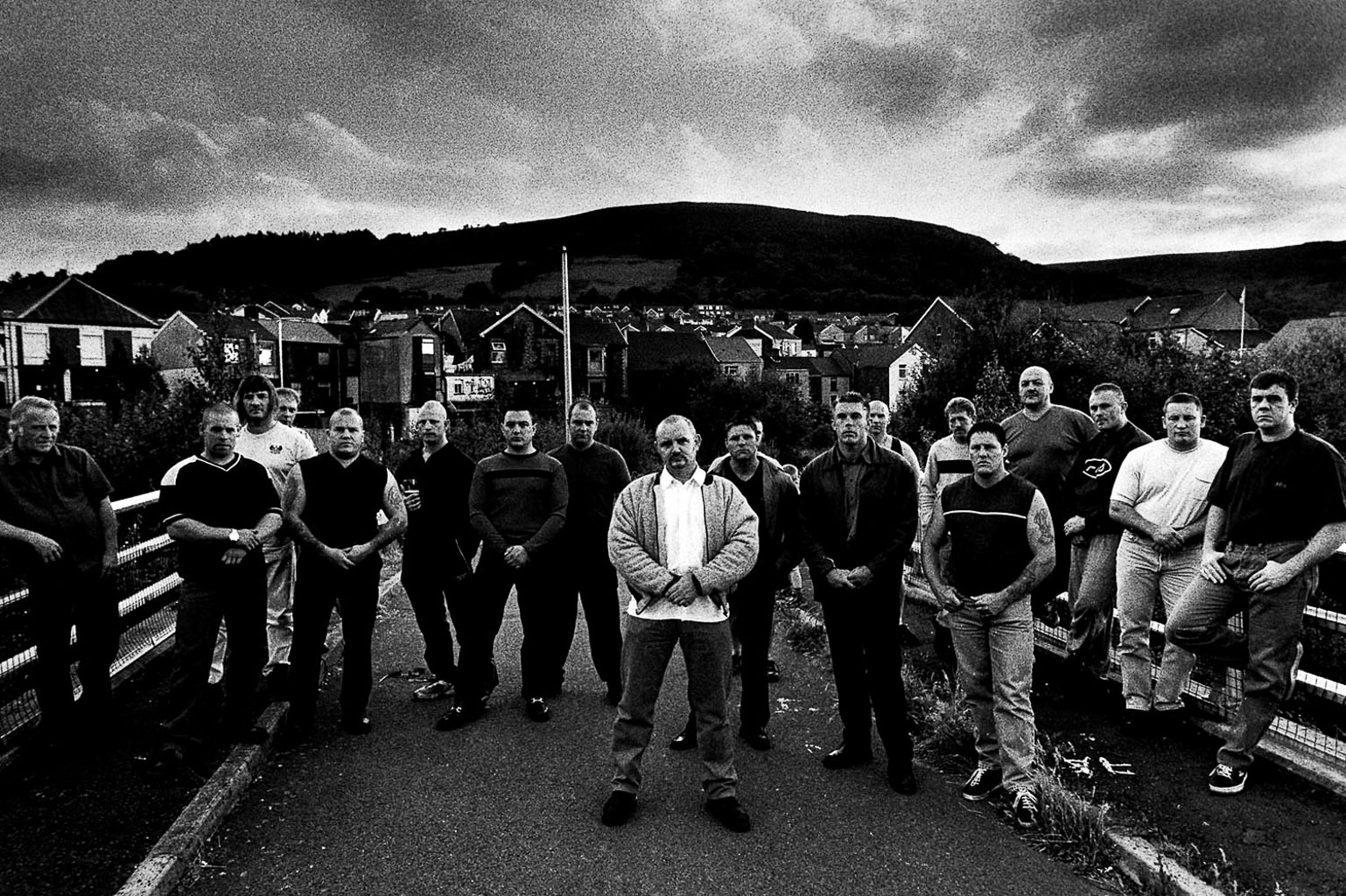 Black and white image of a group of men standing in a row on a road, with houses and hills in the background.