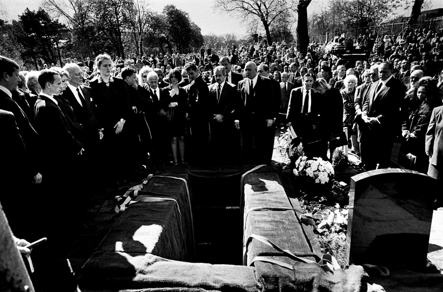 A somber gathering of people in formal attire at a gravesite, surrounded by a crowd, with a partially covered grave and floral arrangements.