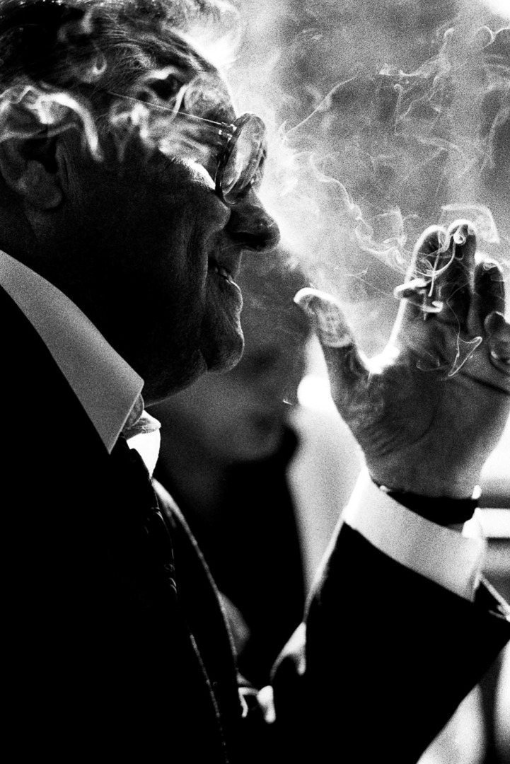 A mature man in a suit smokes a cigarette, creating wisps of smoke around him. He wears glasses and has a thoughtful expression.