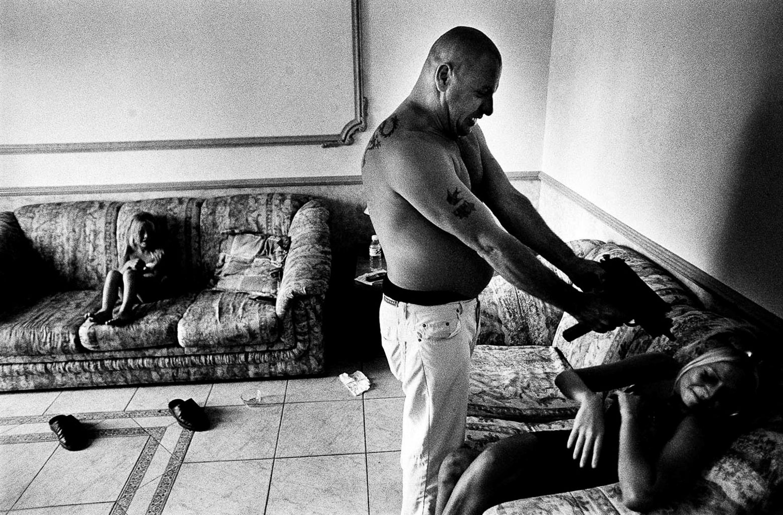 A tense scene showing a man pointing a handgun towards a woman who is lying on a couch, while another child is sitting on a nearby couch, appearing distressed.